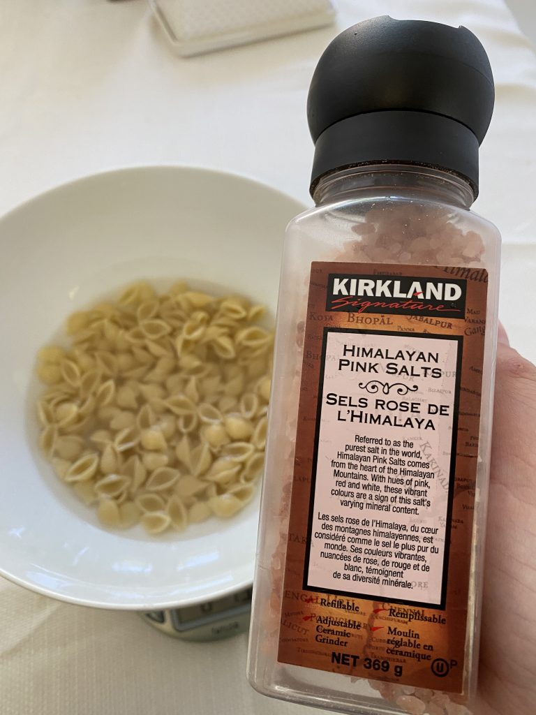 A bowl of noodles and water with a container of Kirkland salt in the forefront of the image.