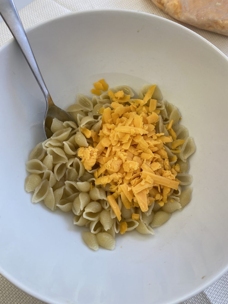 A bowl of cooked noodles topped with freshly grated cheddar cheese with a spoon on the left side of the bowl ready to mix the bowl's contents